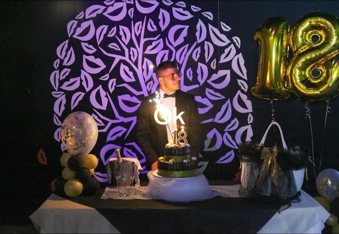 Festeggiato in smoking con scintilla accanto alla torta nera e oro dei 18 anni, davanti all'albero di baci illuminato di viola